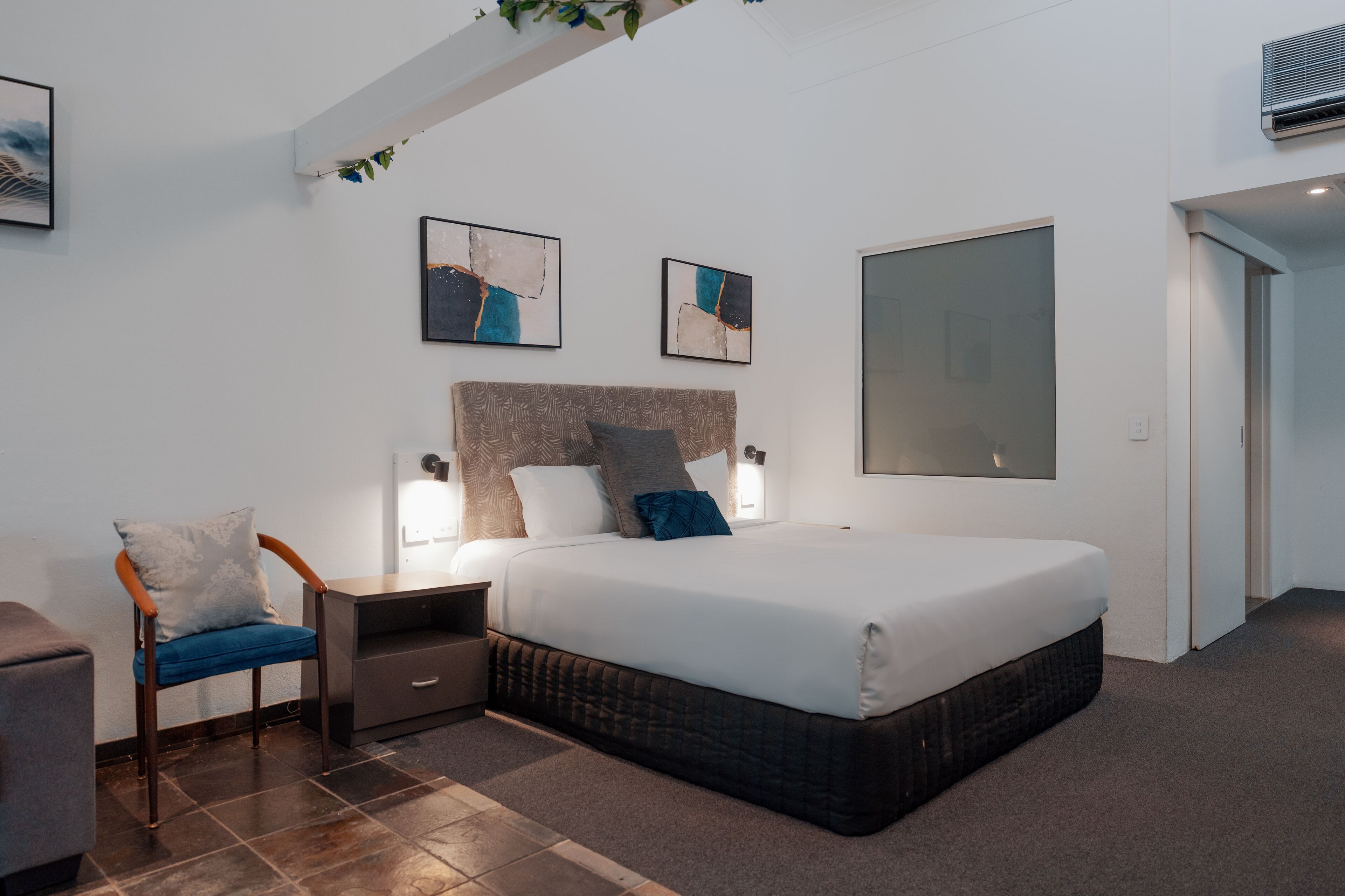 A modern bedroom with a large bed, a nightstand, a chair, and two abstract paintings on the wall.