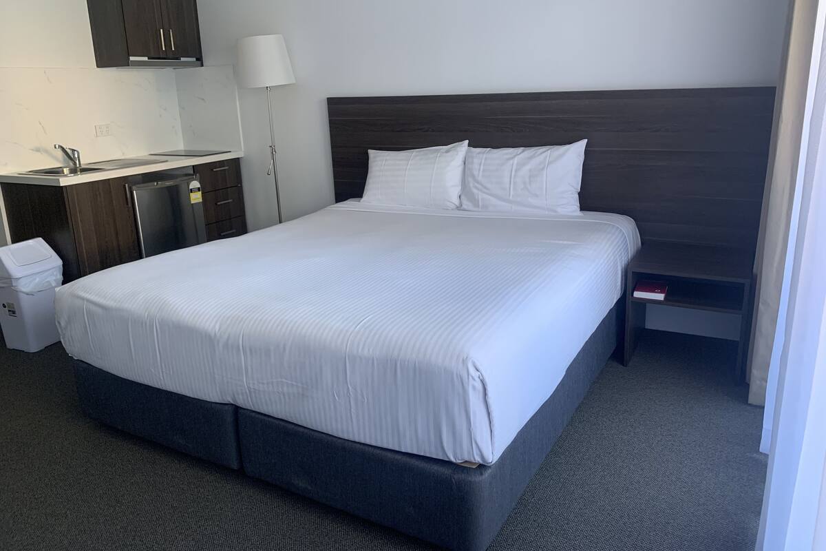 A hotel room with a large bed, a desk with a sink, a trash bin, and a wardrobe.