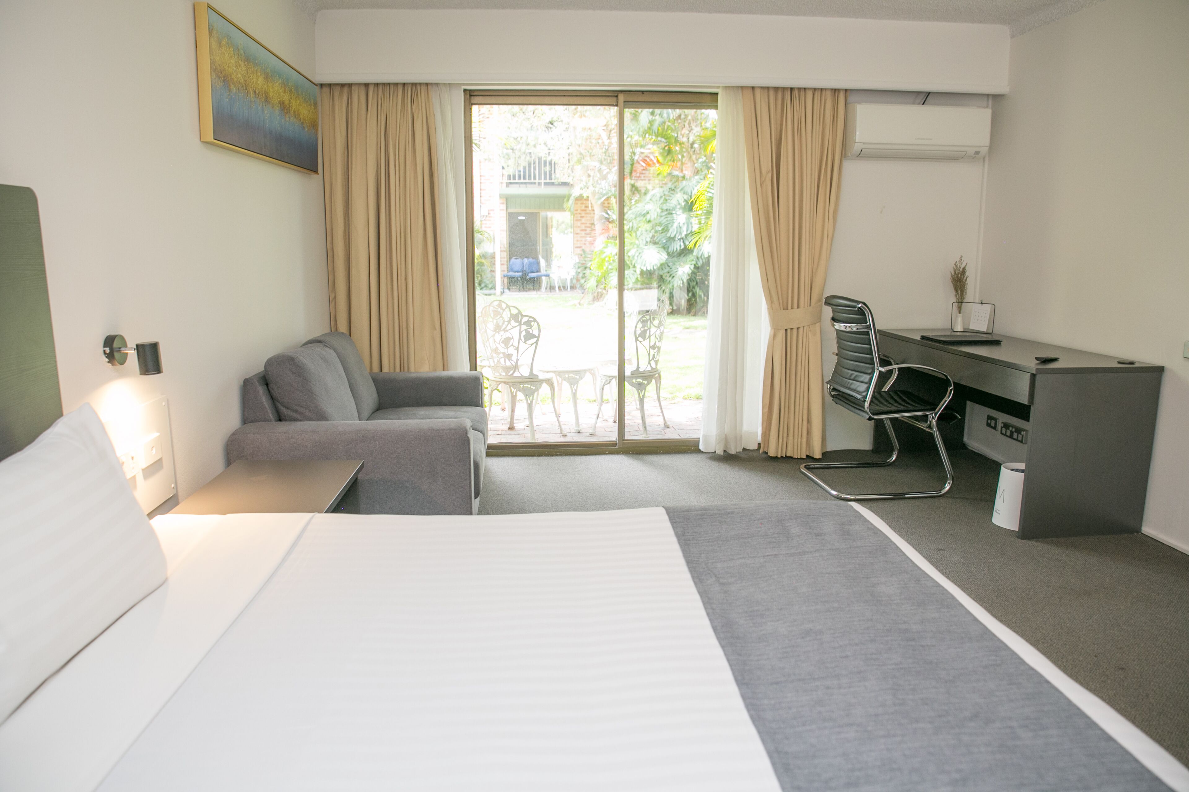 A hotel room with a bed, a sofa, a desk, and a view of the garden through the glass door.