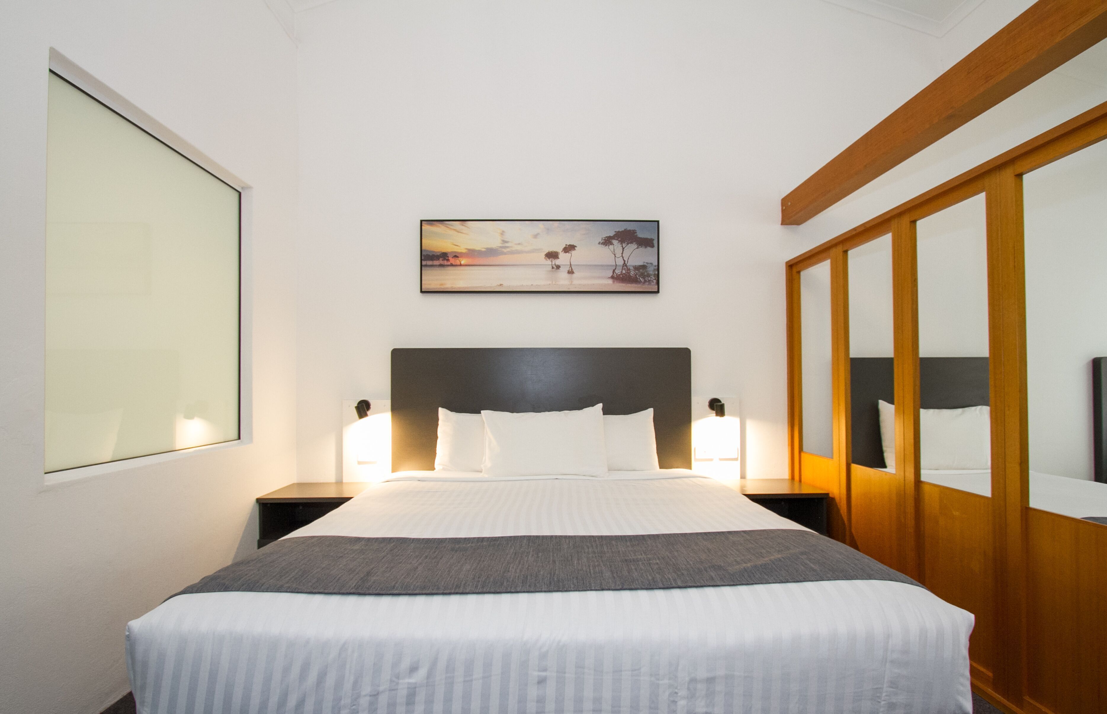 A hotel room with a large bed, two bedside tables, a nightstand, a wooden wardrobe, and a framed picture on the wall.