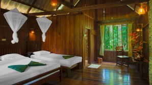 A wooden room with two beds, a window with a view of greenery, and hanging mosquito nets. - Khao Sok Riverside Cottage (Phanom)