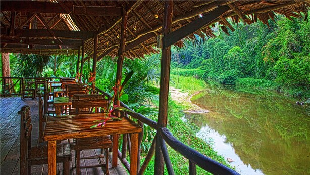 A wooden deck with tables and chairs overlooking a river, surrounded by lush greenery. - Khao Sok Riverside Cottage (Phanom)