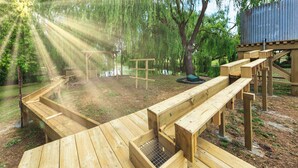 Una terraza de madera con bancos y una pequeña construcción de madera, rodeada de árboles y un cuerpo de agua.