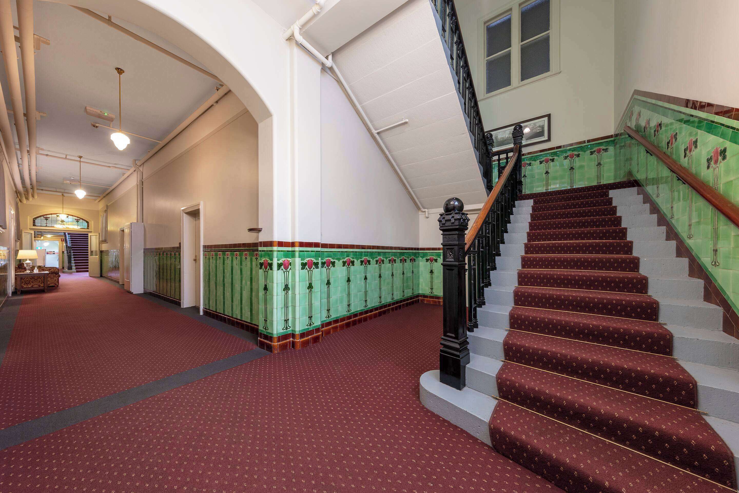 Un grand escalier recouvert d’un tapis rouge mène à un palier, de part et d’autre duquel se trouvent des casiers verts et un plafond blanc.