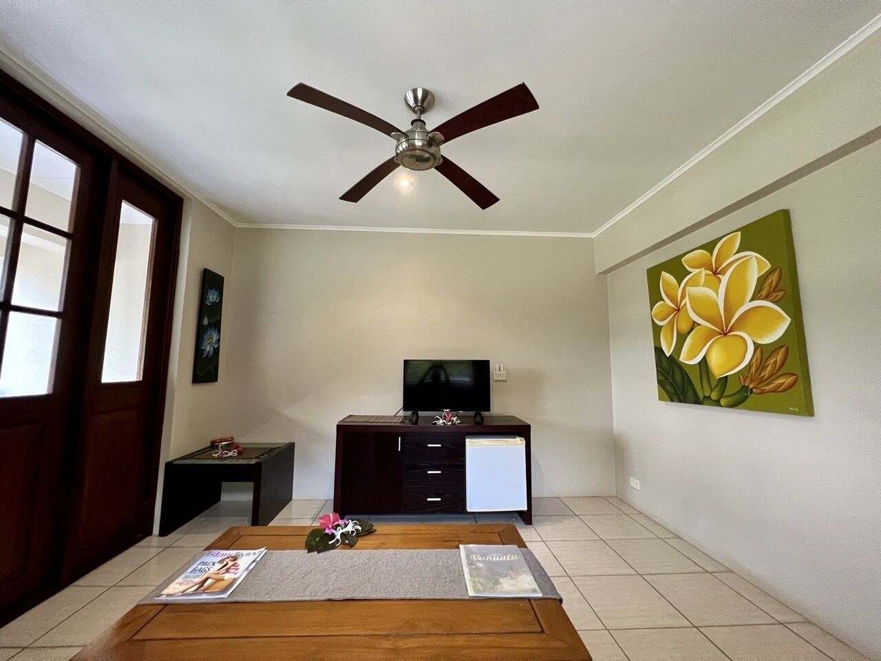 Ein Wohnzimmer mit Deckenventilator, einem Fernseher auf einer Kommode, einem Bild an der Wand und einem Holztisch.