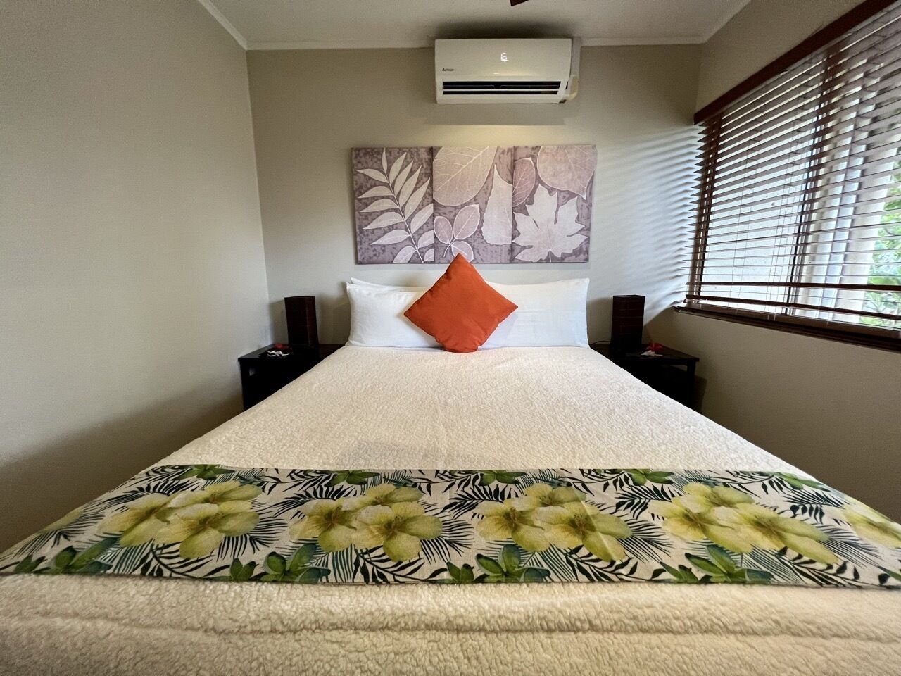 A bedroom with a bed, a wall-mounted air conditioner, a window with blinds, and a floral-patterned bedspread.