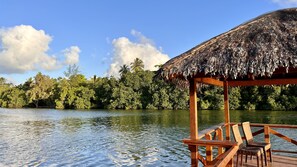 A wooden deck with a thatched roof, overlooking a calm body of water and dense greenery.