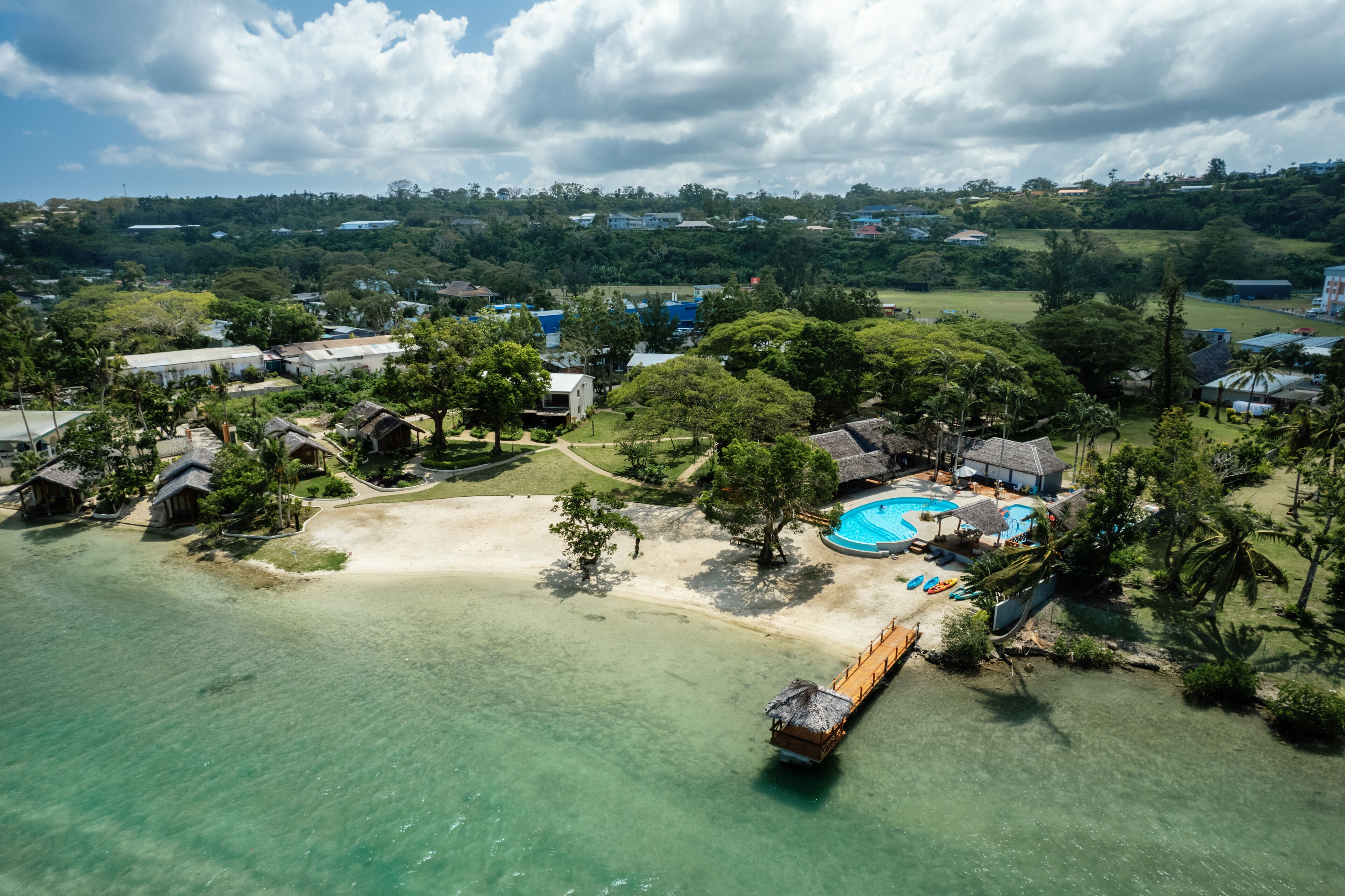 An aerial view of a coastal resort with a swimming pool, beach, and overwater bungalows.