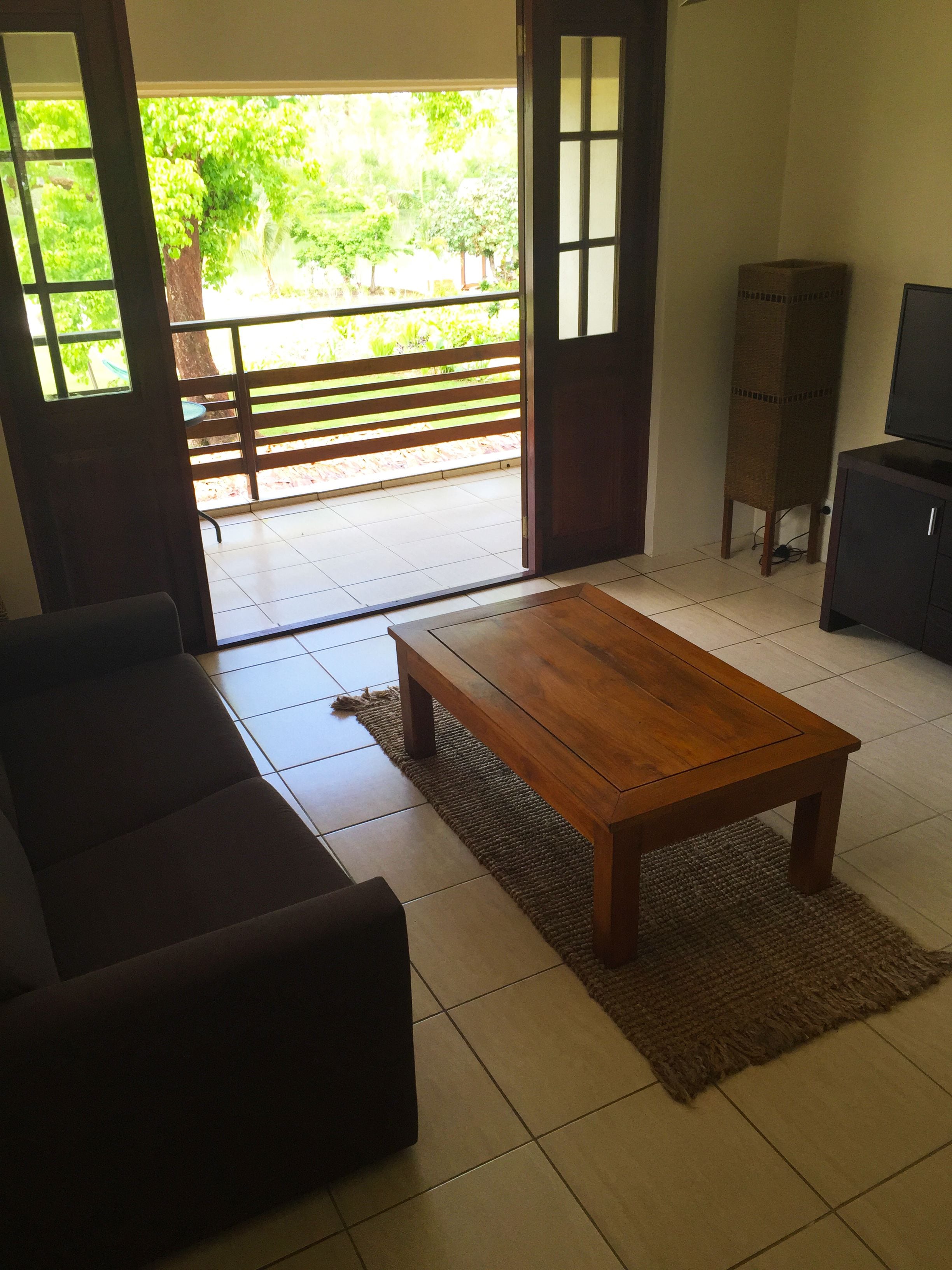 A living room with a wooden coffee table, a dark sofa, a television, and a cabinet. There is a sliding glass door leading to a balcony with a view of greenery.