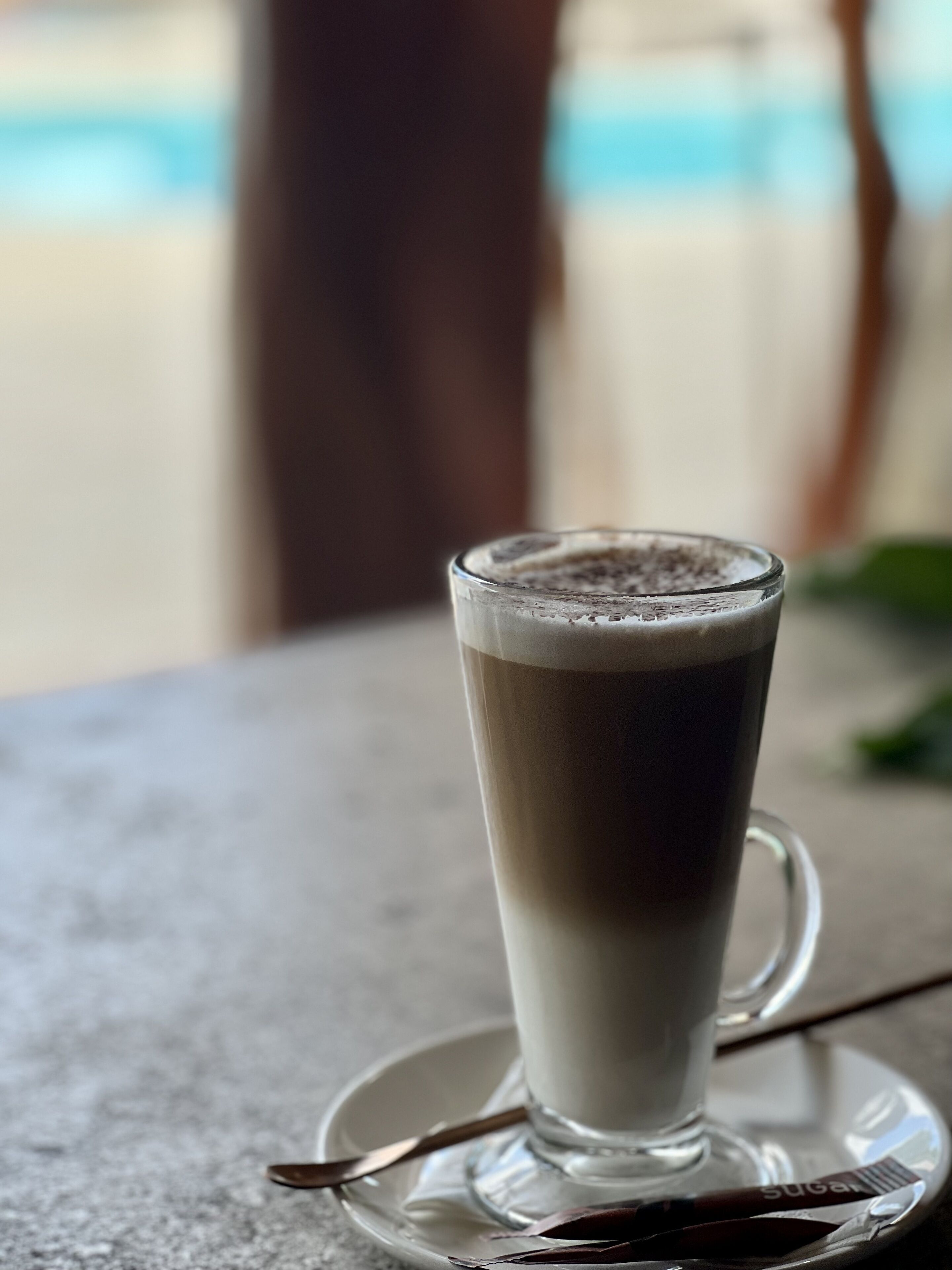 A tall glass of layered beverage on a saucer with a spoon, placed on a table.