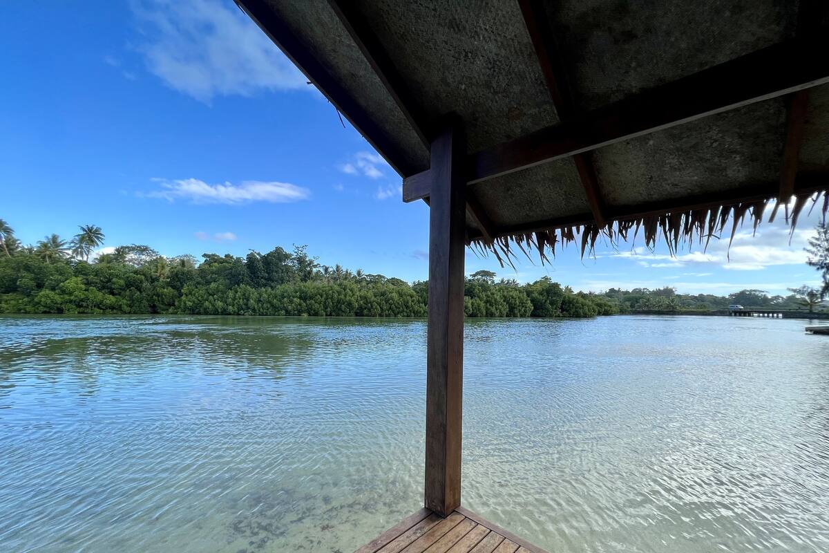 Eine Holzterrasse mit Blick auf ruhiges Wasser, darüber ein Reetdach.