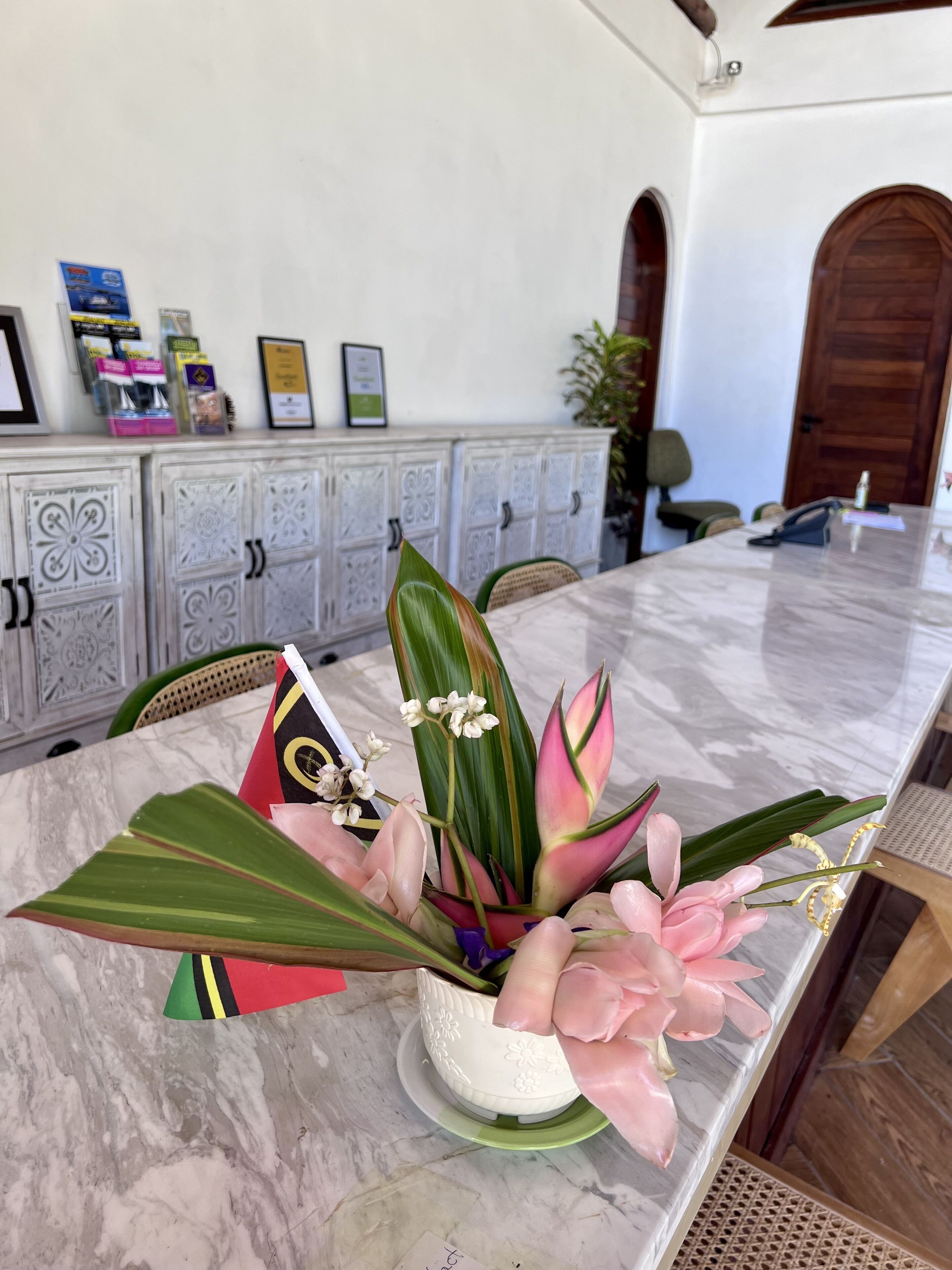 Una mesa de mármol con un centro de mesa floral, un aparador blanco con puertas decorativas y diplomas enmarcados en la pared.