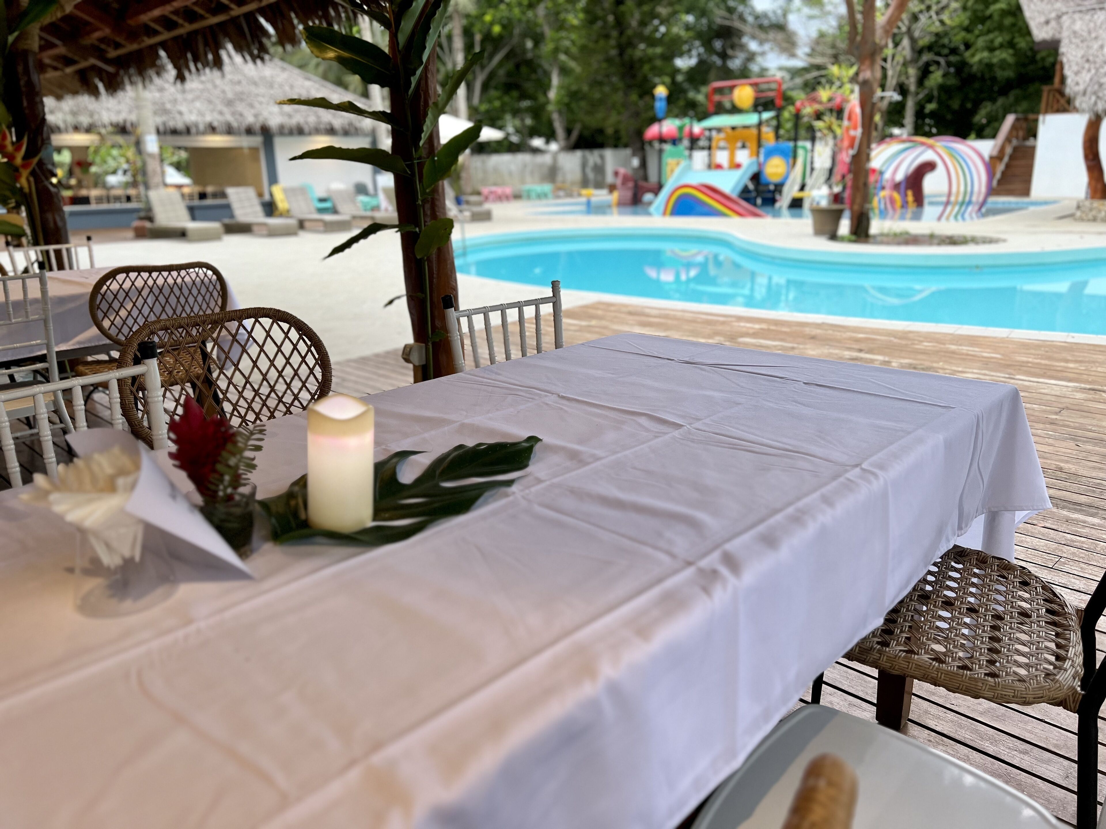 Un área de comedor al aire libre con una mesa dispuesta para dos, con vista a una piscina y una zona de juegos infantiles.