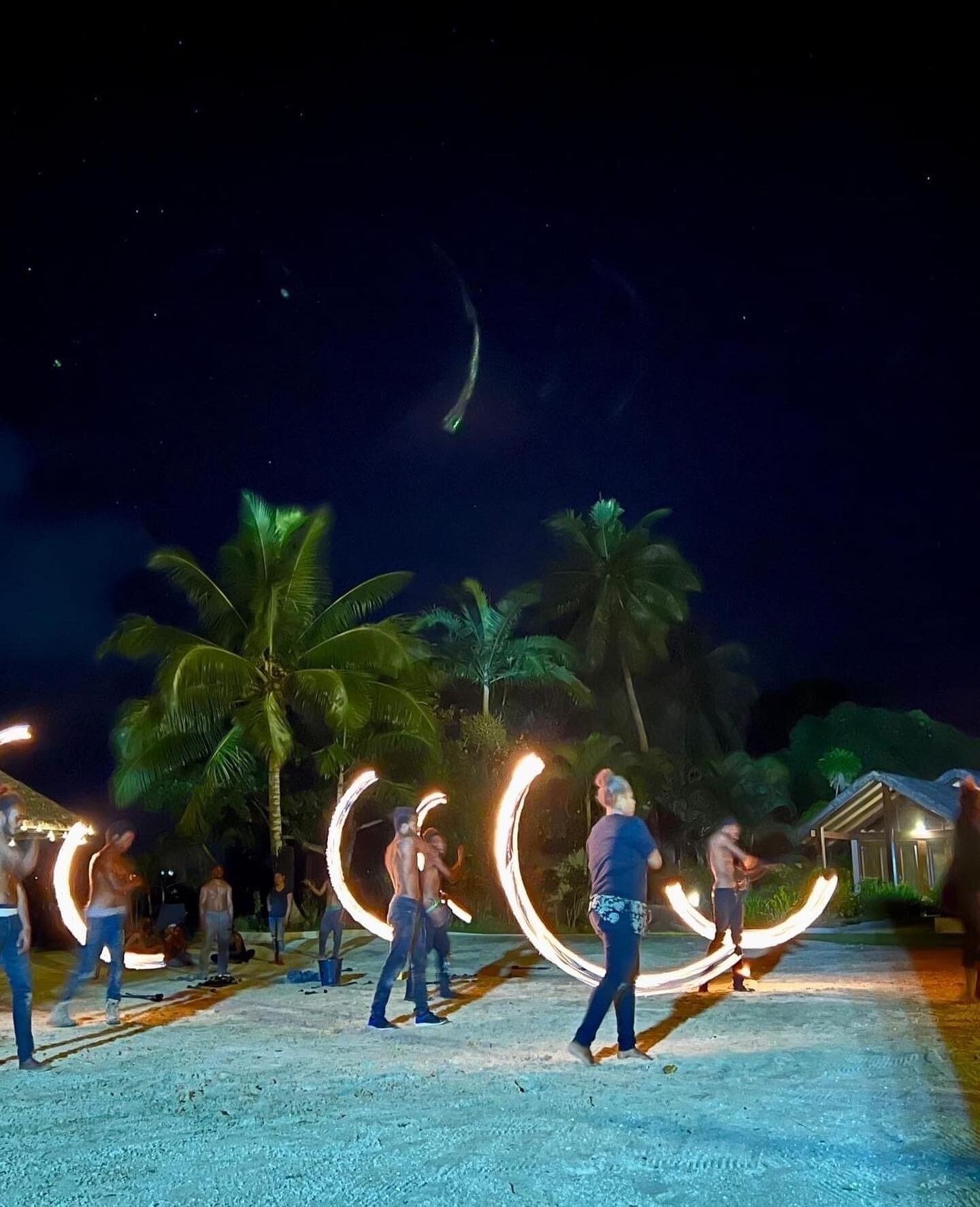 A nighttime scene with people performing fire dancing on a sandy area surrounded by palm trees.