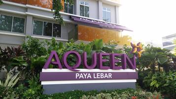 Señalización del Hotel Aqueen en Paya Lebar, rodeado de exuberante vegetación.