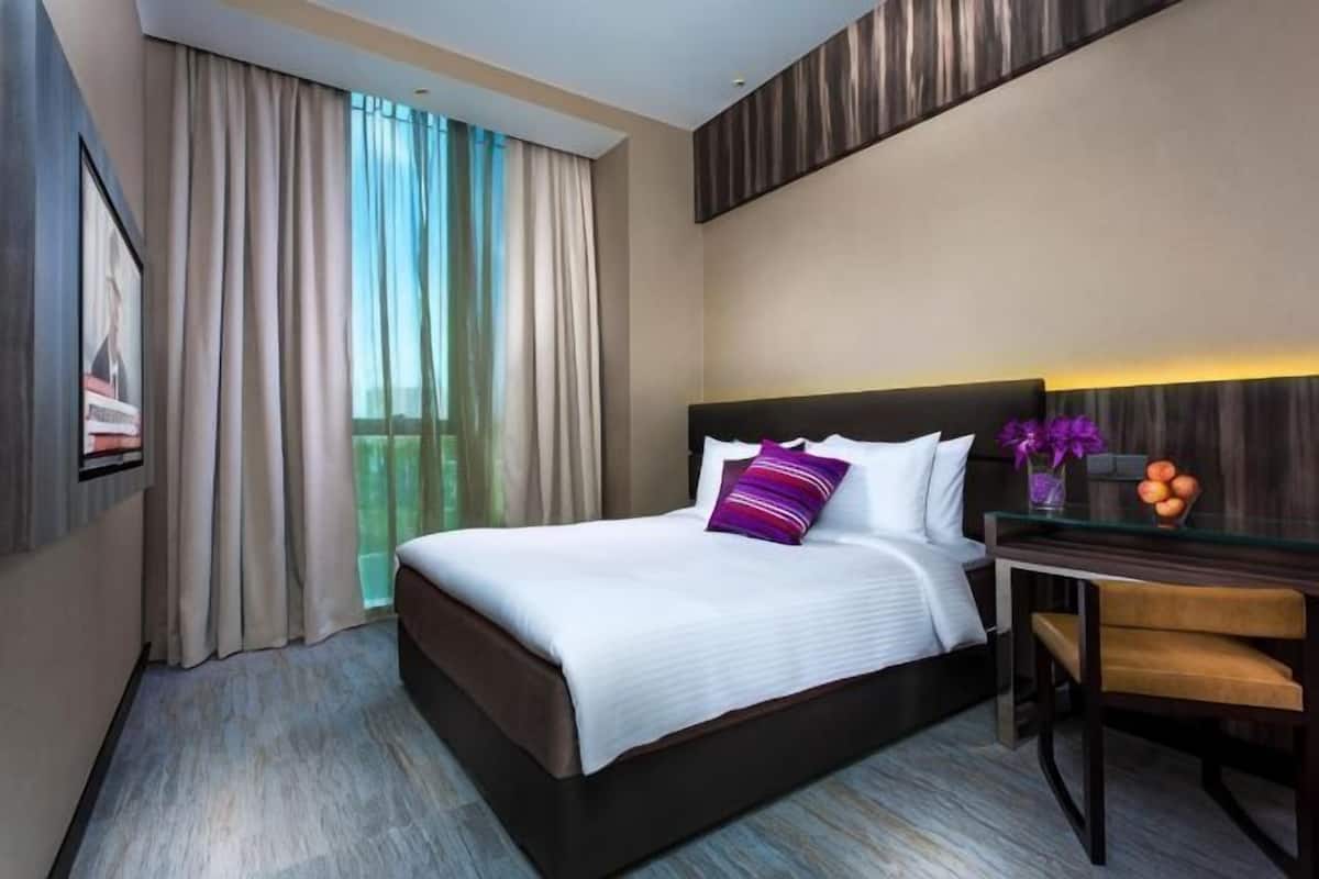 Habitació d'hotel amb llit, escriptori, cadira, finestra amb cortines i televisió penjada a la paret.