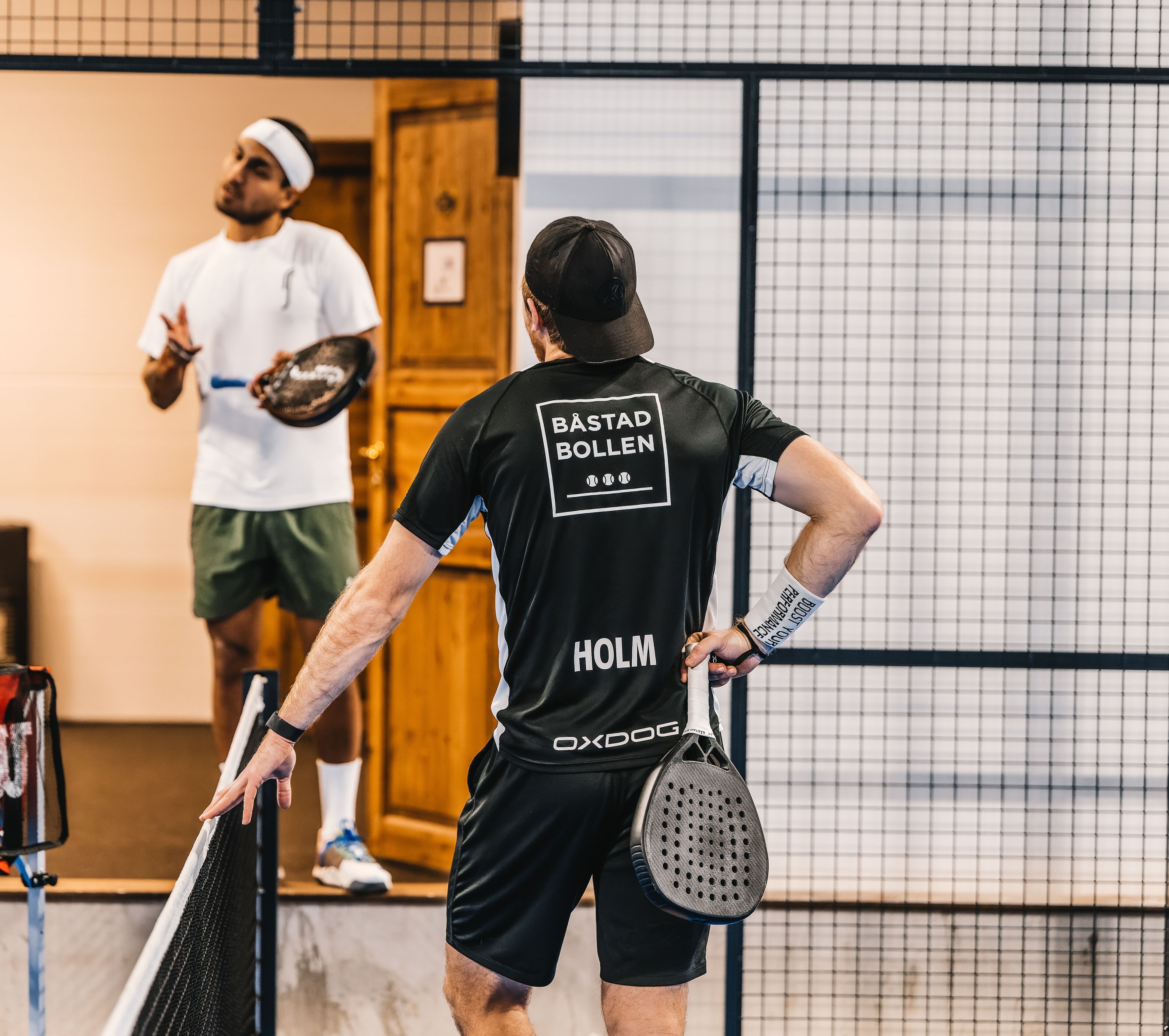 Ein Tennisspieler mit schwarzem Trikot, auf dessen Rückseite "BÄSTAD BOLLEN" und "HOLM" stehen, hält einen Tennisracket und steht auf einem Tennisplatz mit Netz im Hintergrund.