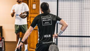 Ein Tennisspieler mit schwarzem Trikot, auf dessen Rückseite "BÄSTAD BOLLEN" und "HOLM" stehen, hält einen Tennisracket und steht auf einem Tennisplatz mit Netz im Hintergrund.