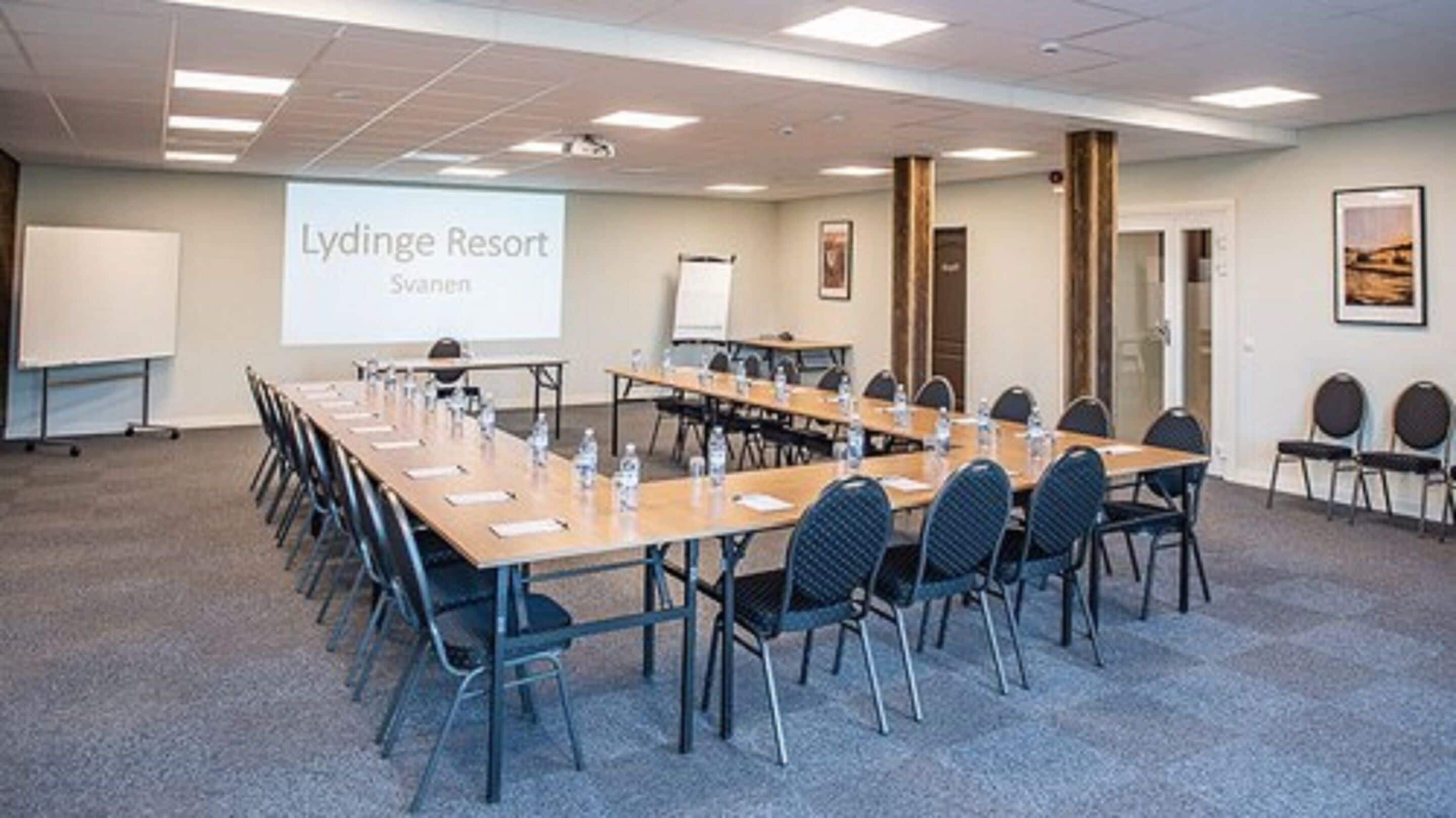 Un salón de conferencias en Lydinge Resort con una larga mesa, sillas, una pantalla de proyector y una pizarra.