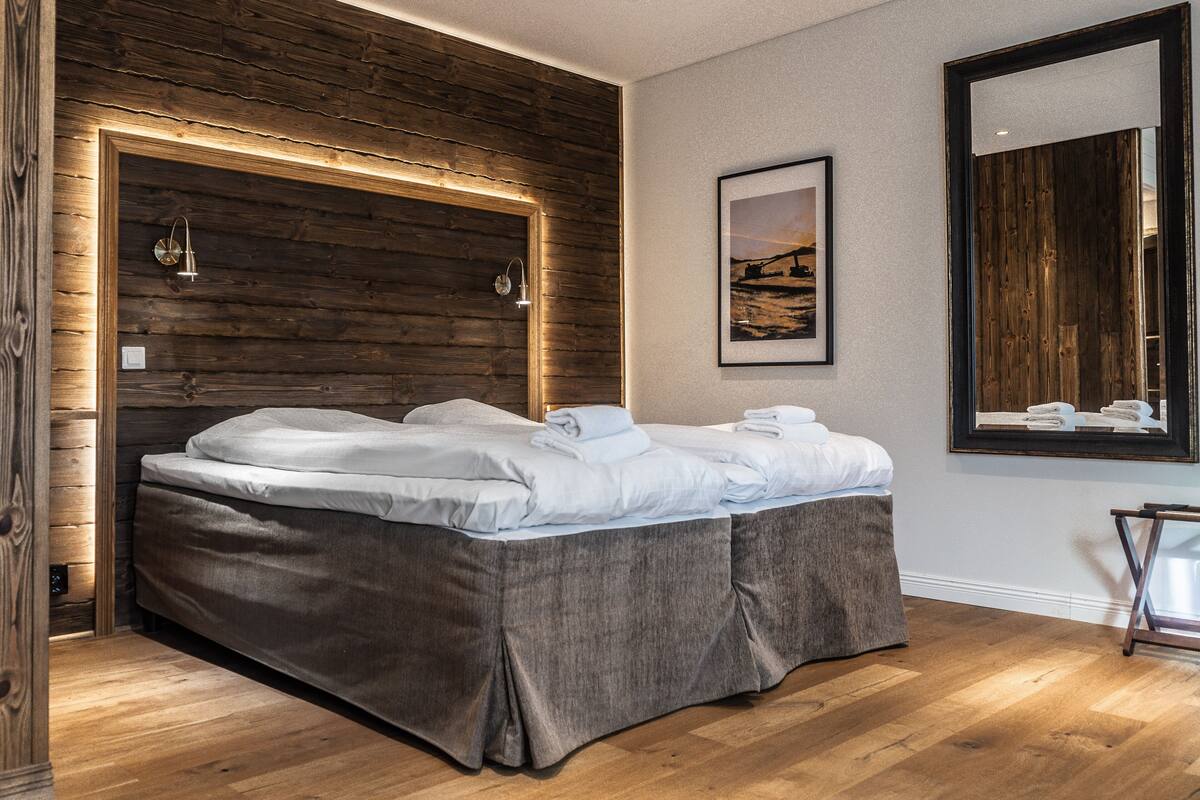 Une chambre moderne avec une tête de lit en bois, un grand lit recouvert de draps blancs soigneusement pliés, un tableau représentant un paysage encadré accroché au mur et un miroir qui reflète la pièce.