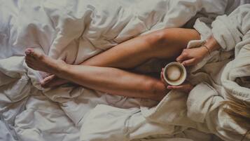 Une personne allongée sur un lit aux draps blancs, tenant une tasse de café.