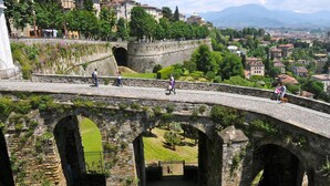 An ancient stone bridge with multiple arches, surrounded by lush greenery and historic buildings in the background. - Al San Lorenzo (Bergamo)