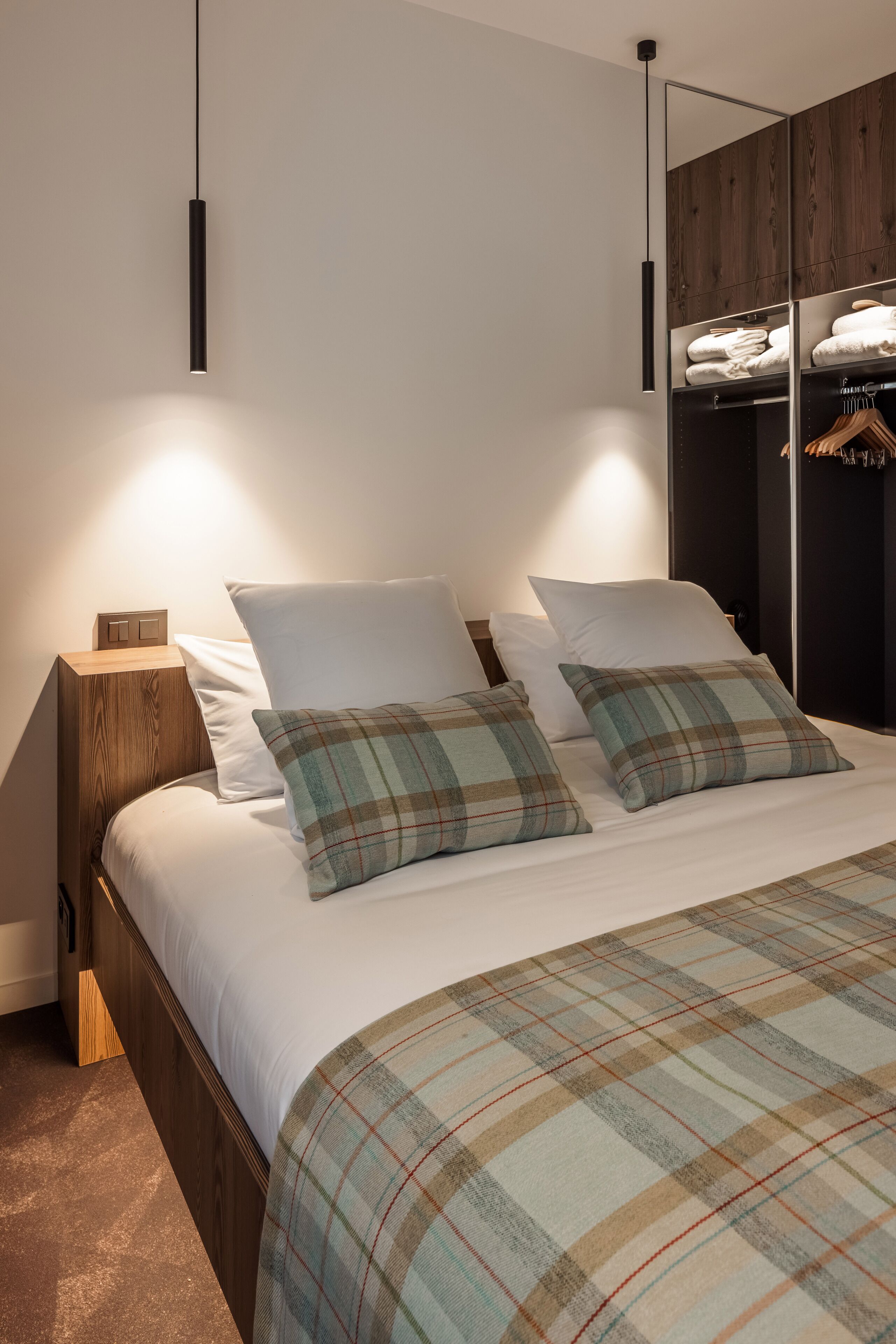 A modern bedroom with a wooden headboard, a large bed with plaid bedding, a nightstand with a lamp, and a wardrobe with hanging clothes.