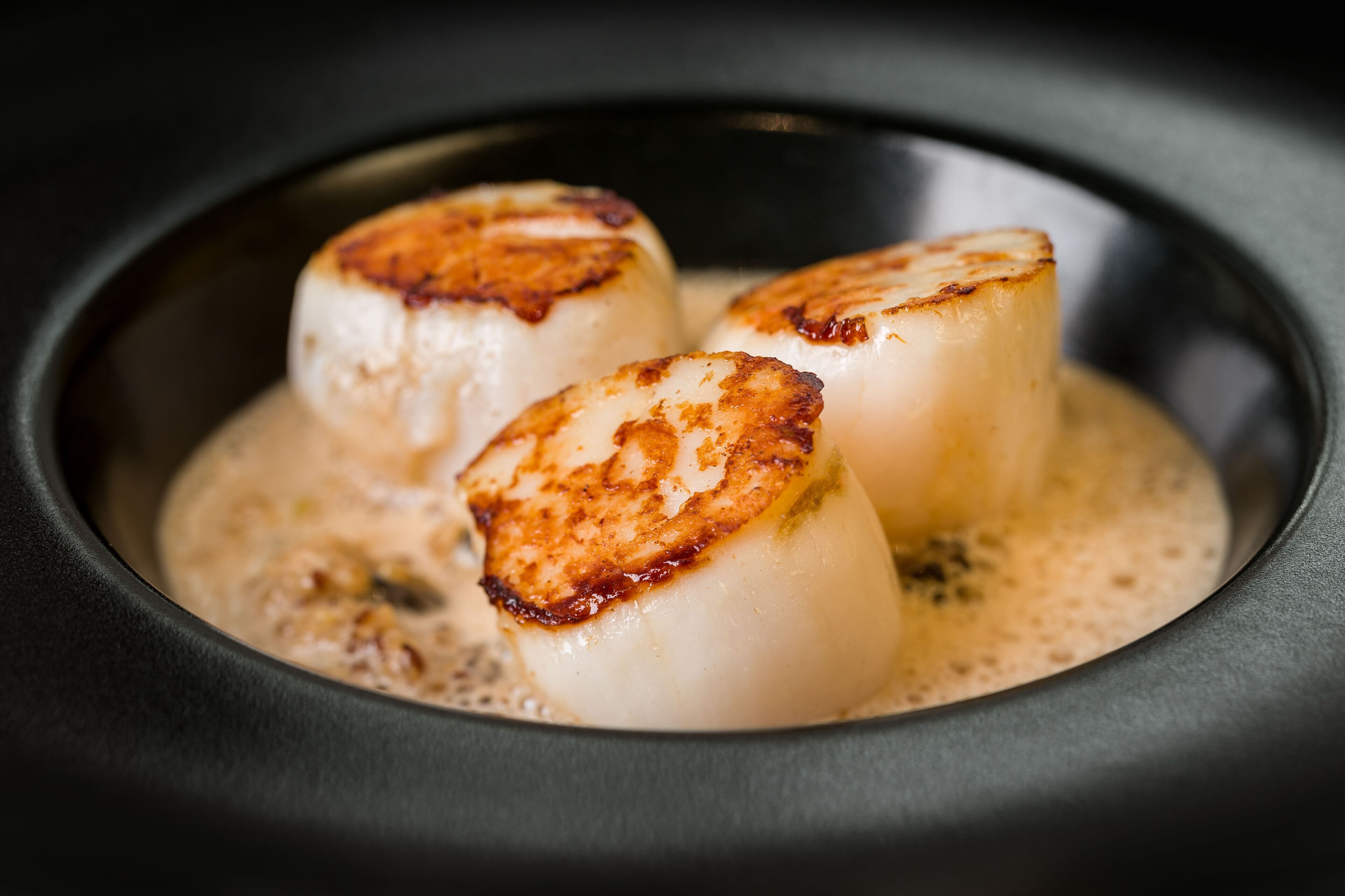 Three scallops in a black bowl with a creamy sauce.