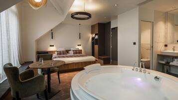 Una habitación de hotel moderna con una cama grande, un área de descanso con una mesa y sillas, una bañera amplia y un baño con espejo y lavamanos.