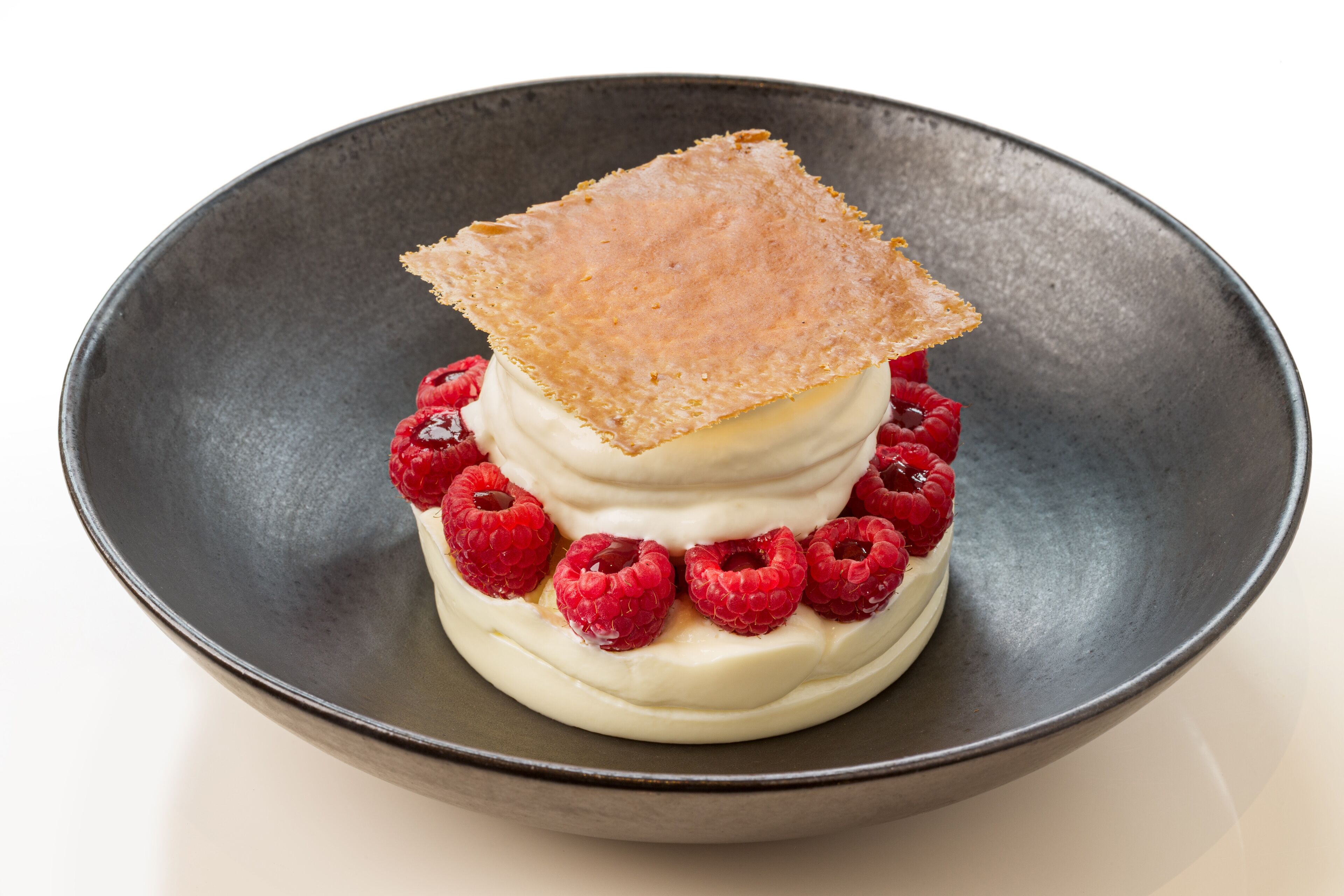 Un postre con una porción cuadrada encima, rodeado de frambuesas, en un bol oscuro.