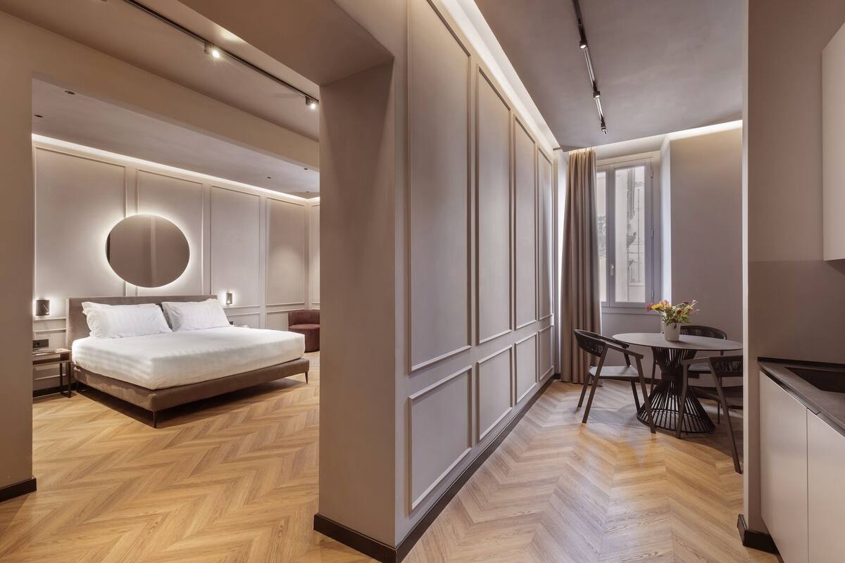 Una habitación de hotel moderna con una cama grande, un espejo redondo, una mesa de comedor con sillas y vistas a la ciudad desde la ventana.