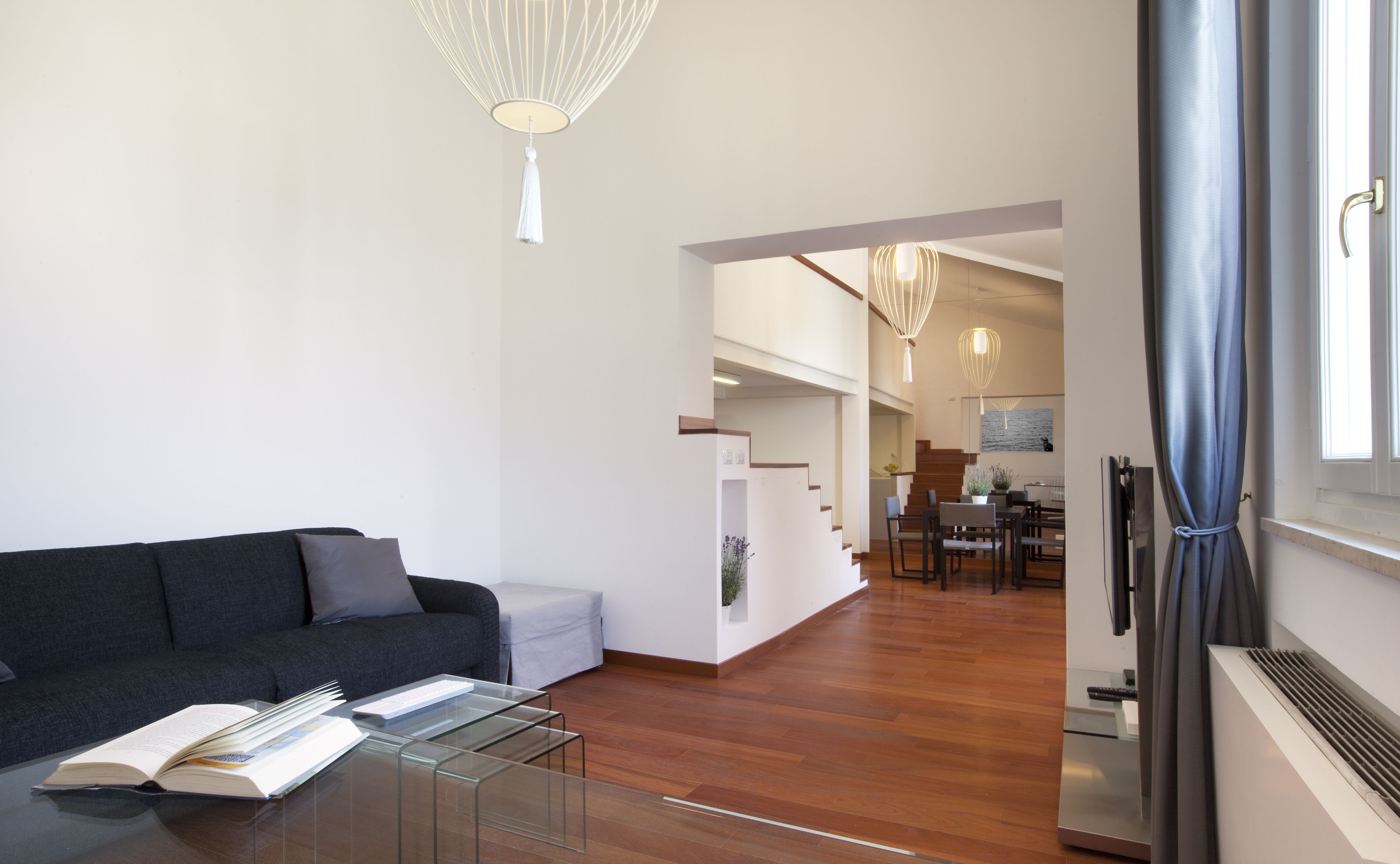 A modern living room with a dark sofa, a glass coffee table, and a dining area with wooden flooring and a staircase leading to an upper level.