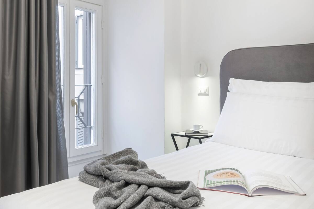 Una habitación de hotel con una cama, una ventana con cortinas, una mesita de noche con un libro y una taza, y una lámpara fijada a la pared.