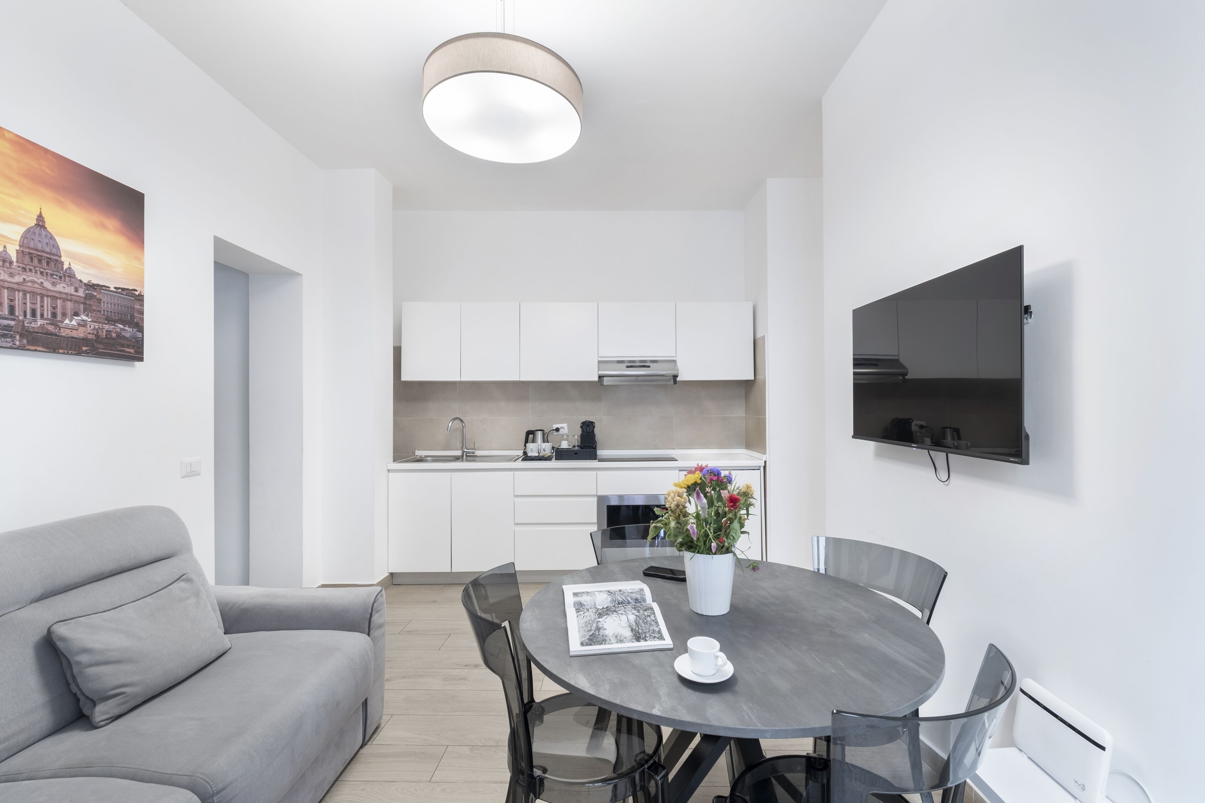 A modern living room with a grey sofa, a round dining table with chairs, a flat-screen TV mounted on the wall, and a kitchen in the background.