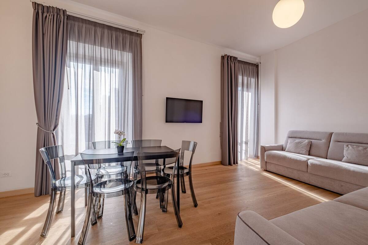 Una sala de estar moderna con una mesa redonda de vidrio, sillas transparentes, un sofá color beige y un televisor de pantalla plana montado en la pared.