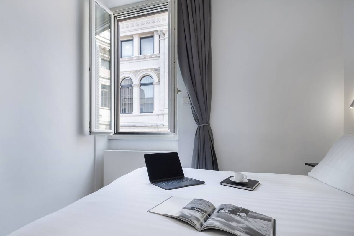 Una habitación de hotel con una cama, una computadora portátil, una taza y una revista abierta.