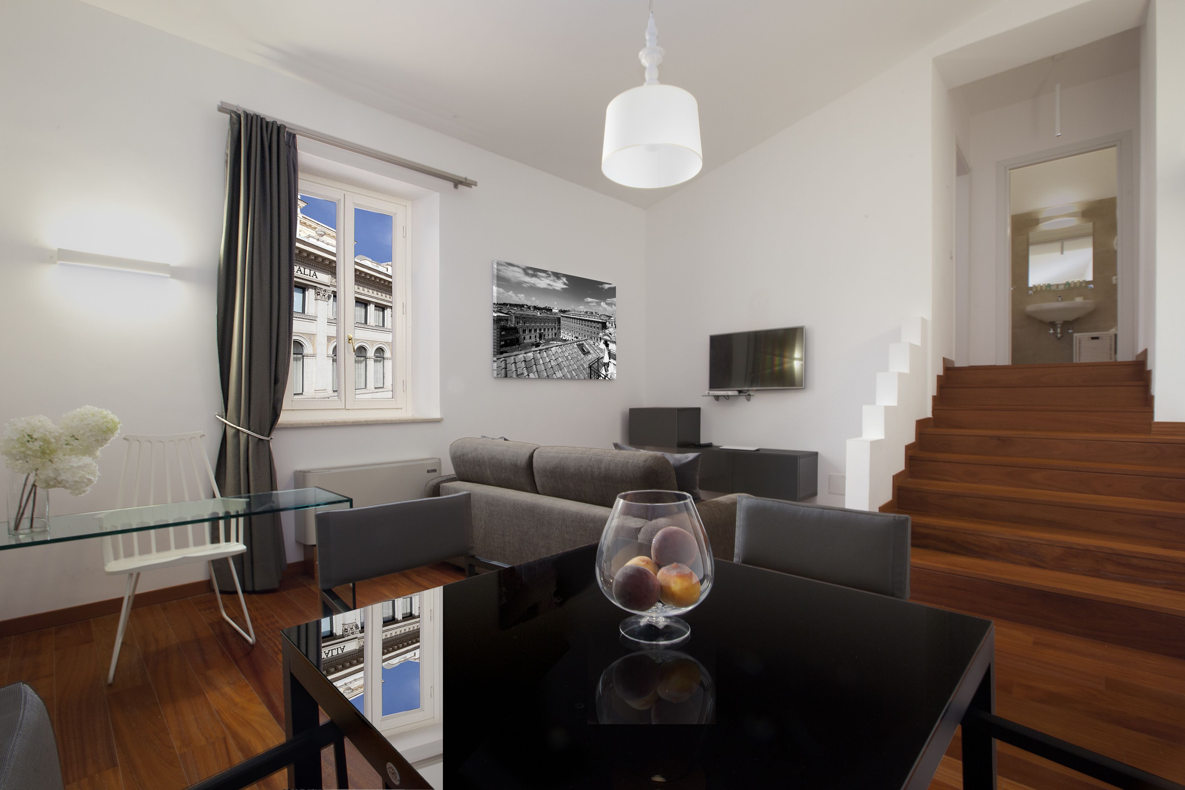 Een moderne woonkamer met een glazen salontafel, een grijze bank, een zwarte tv-meubel en een trap naar een verdieping daarboven.