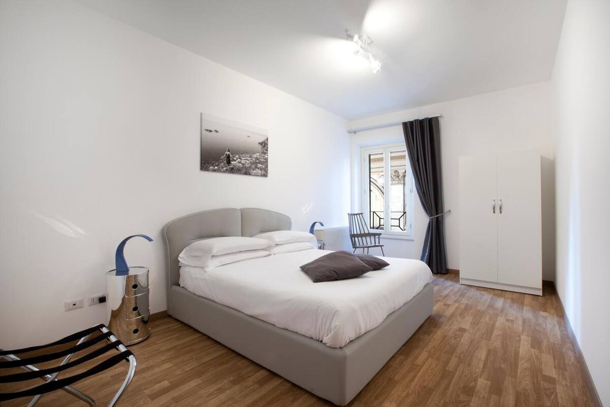 Un dormitorio moderno con una cama grande, una mesita de noche, una silla, un armario y una ventana con cortinas.