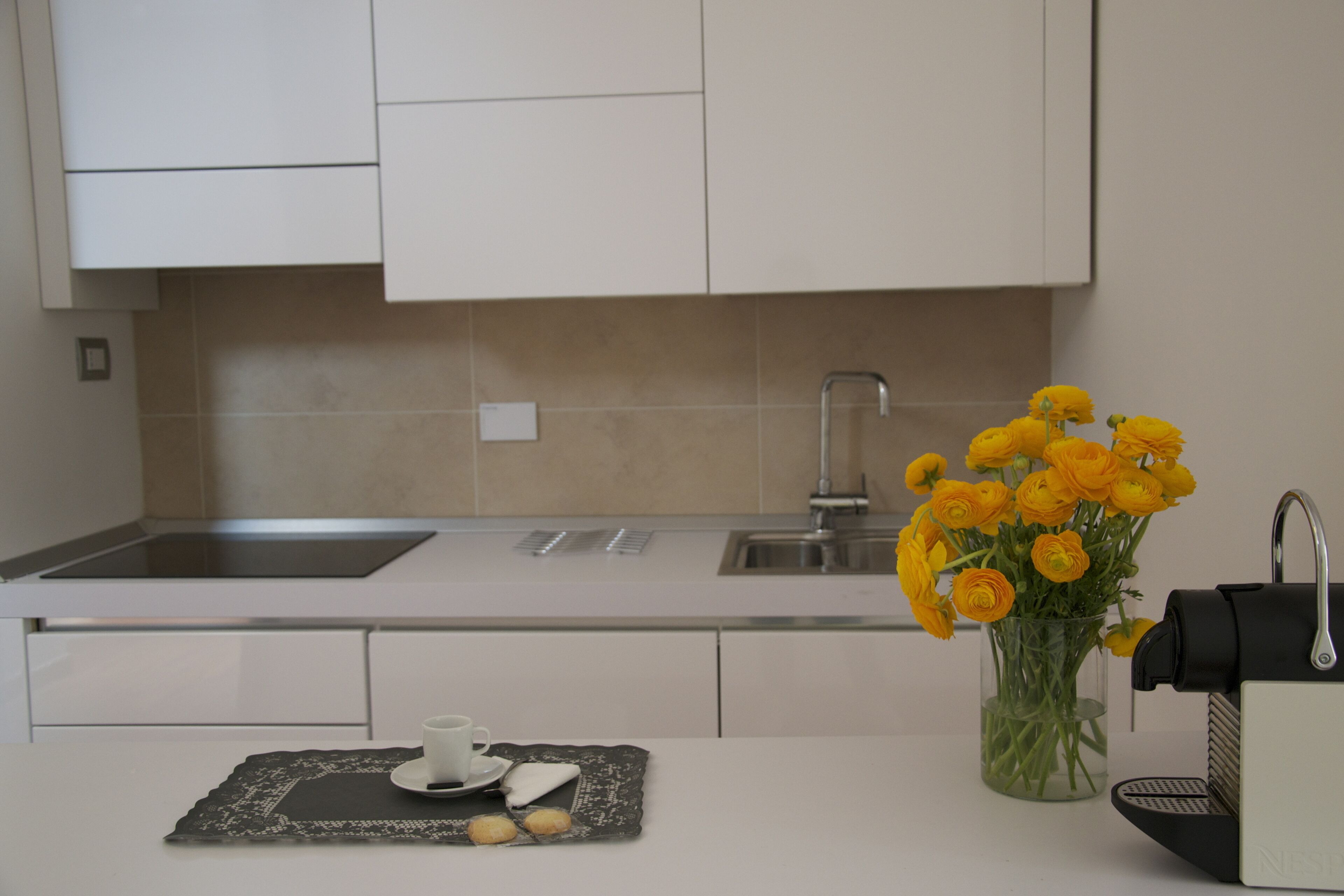Una cucina moderna con mobili bianchi, un lavello in acciaio inossidabile e un vaso di fiori gialli sul piano di lavoro.