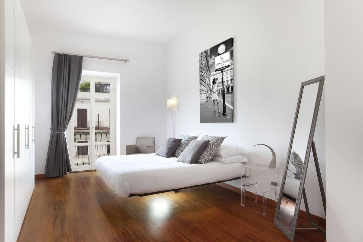 Un dormitorio moderno con una cama grande, una mesita de noche, una lámpara y una obra de arte con un paisaje urbano en la pared.