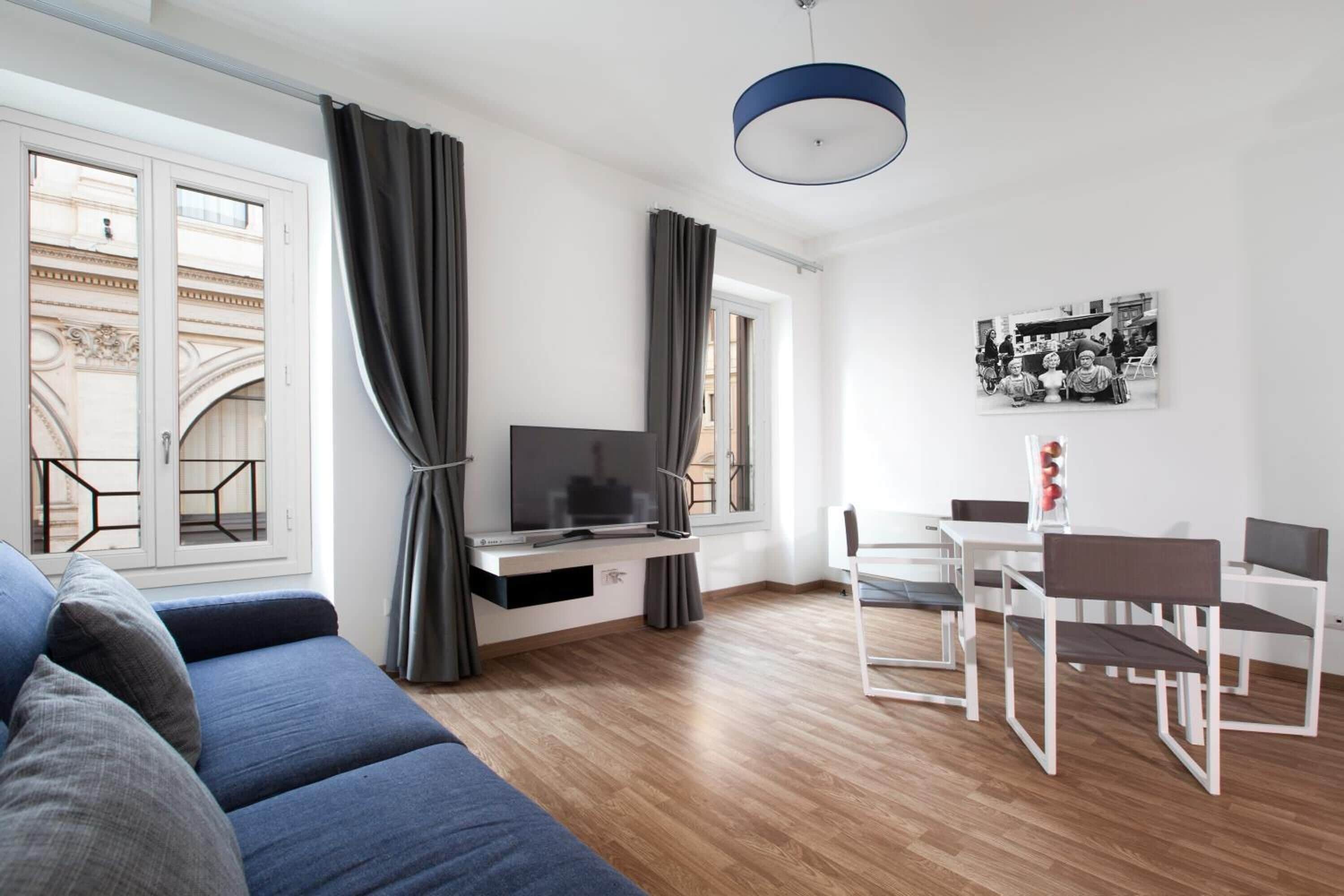 A modern living room with a sofa, TV, dining table, chairs, and a large window with curtains.