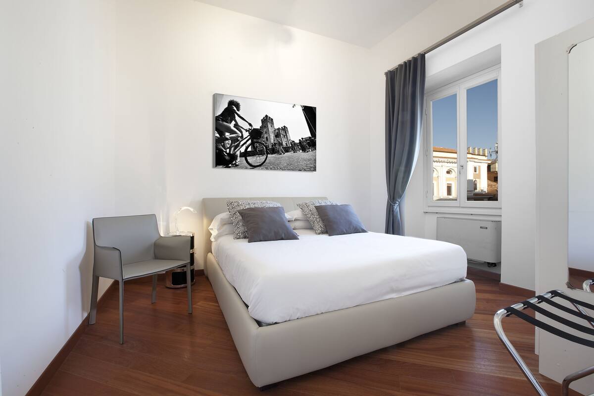 Un dormitorio moderno con una cama grande, una silla, una mesita de noche y un ventanal con cortinas.