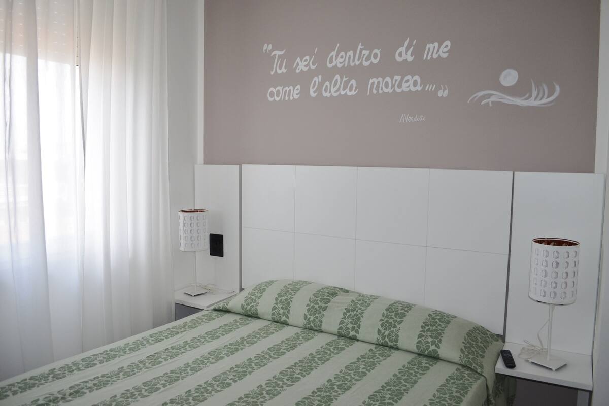 A hotel room with a bed, a bedside table, a lamp, and a wall with a quote.