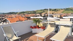 A rooftop terrace with two lounge chairs, a potted plant with red flowers, and a view of a town with red-roofed buildings and green hills in the distance. - Hotel Sette Note (Silvi)