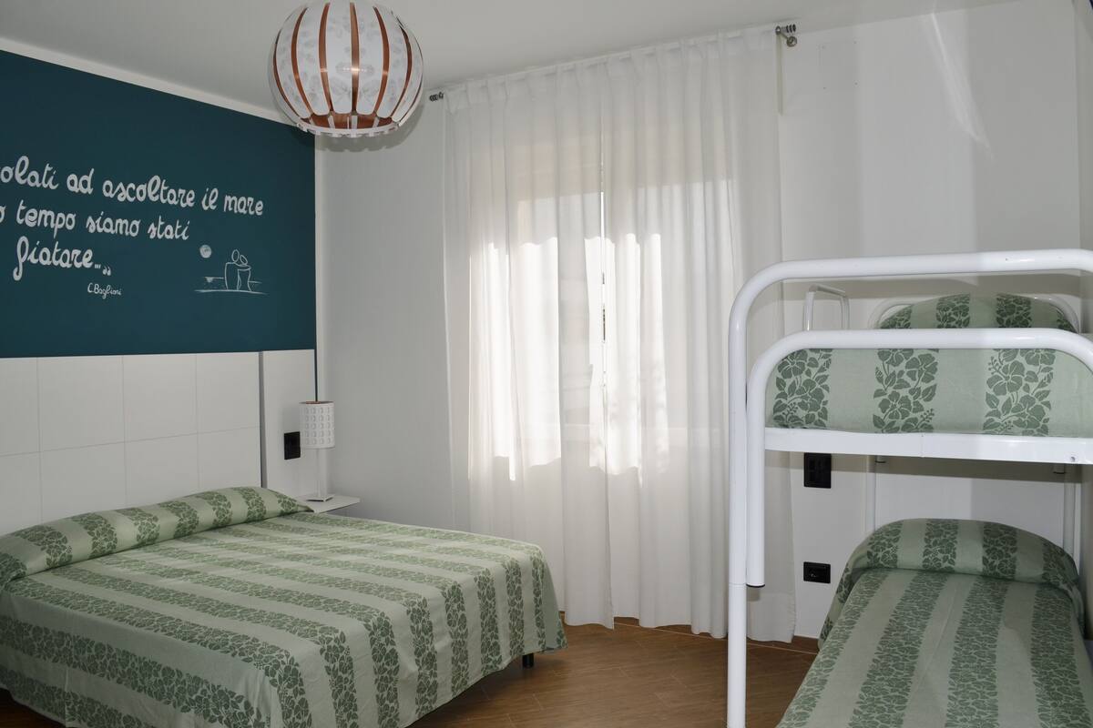 A room with two single beds, a bunk bed, a white ceiling fan, a wall with a quote, and a window with sheer curtains.