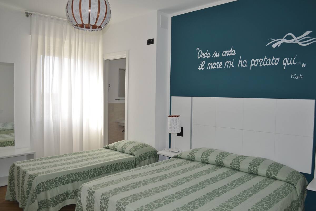 A hotel room with two beds, a window with sheer curtains, a wall quote, and a bedside lamp.