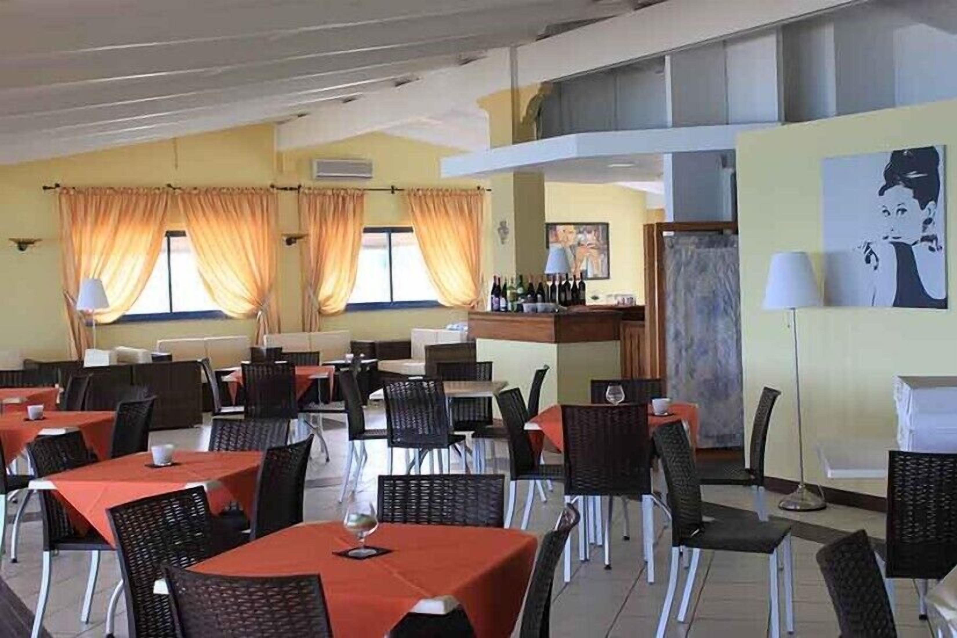 A restaurant with tables covered in red tablecloths and black chairs, a bar area with bottles, and a painting on the wall.