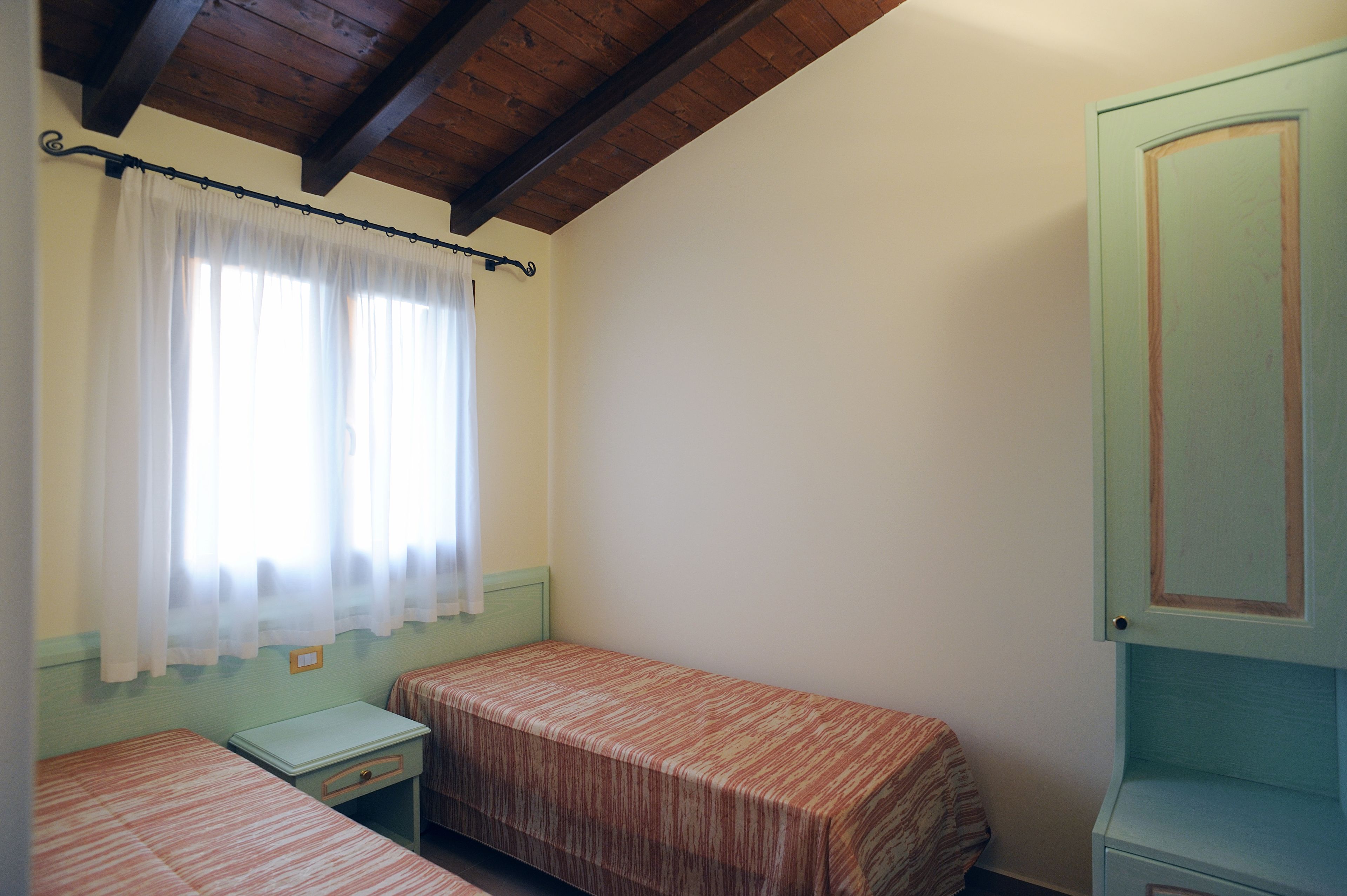 Une chambre avec deux lits simples, une petite table, une armoire et une fenêtre avec des rideaux.