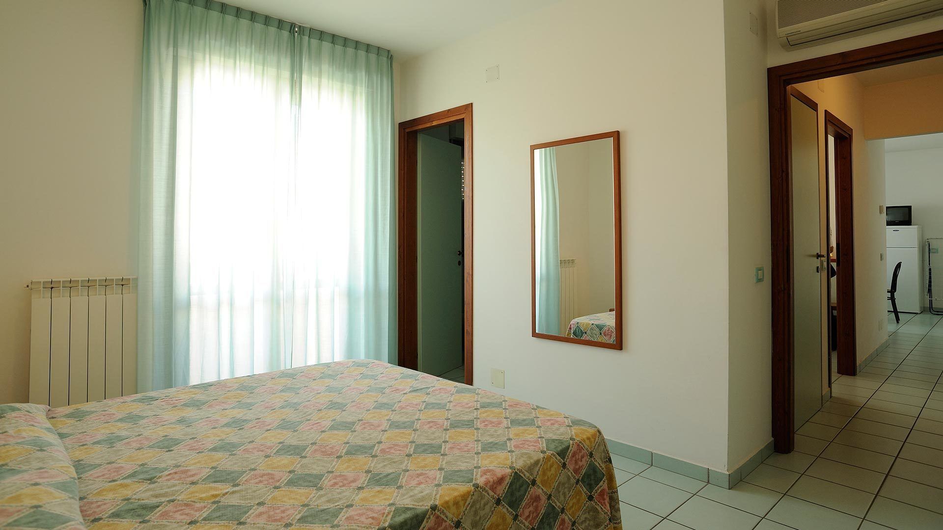 Una camera da letto con un letto, un termosifone, una finestra con tende, uno specchio e un ingresso verso una piccola cucina.