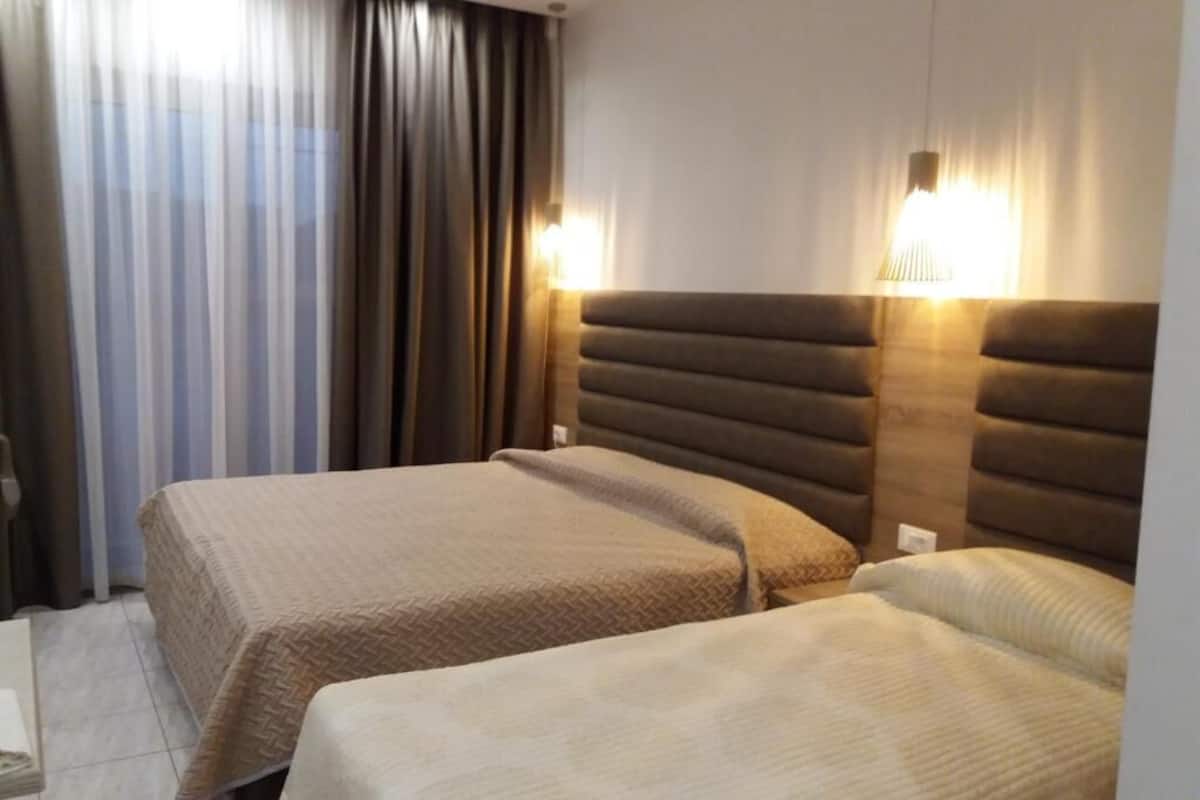 Habitación de hotel con dos camas, un ventanal grande con cortinas y lámparas fijadas a la pared.